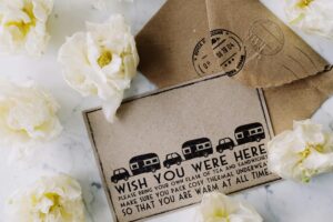 Vintage postcard reading “Wish You Were Here” surrounded by white flowers and brown envelopes, evoking nostalgic travel vibes.