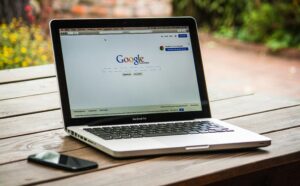 Laptop displaying Google search page on a wooden table outdoors with a smartphone beside it