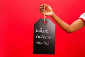 A hand with red-painted nails holds a black tag against a bright red background, with the handwritten message “Where customers matter.”