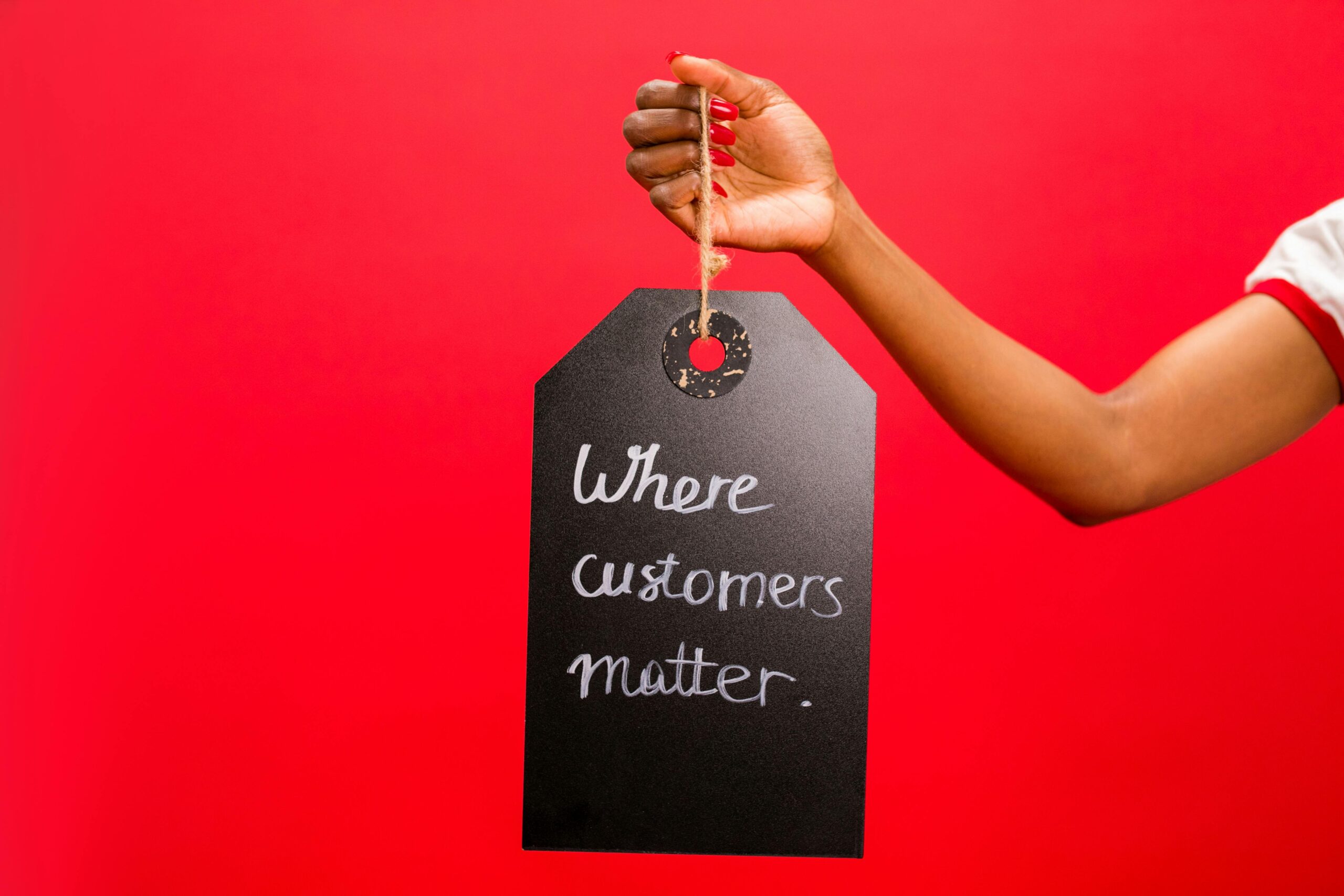 A hand with red-painted nails holds a black tag against a bright red background, with the handwritten message “Where customers matter.”